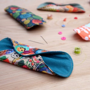 May include: A teal fabric pouch with a colourful print of Russian nesting dolls. The pouch is folded in half and has a button closure. The pouch is sitting on a wooden surface with sewing supplies.