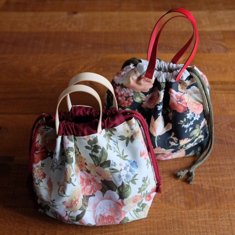 May include: Two floral drawstring bags with different patterns. One bag has a dark background with white and pink flowers. The other bag has a light background with pink, red, and blue flowers. Both bags have burgundy fabric lining and wooden handles.