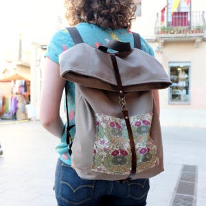 Könnte beinhalten: Ein brauner Canvas-Rucksack mit einer Tasche mit Blumenmuster und braunen Lederriemen. Der Rucksack hat eine umklappbare Klappe mit einem Schnallenverschluss.
