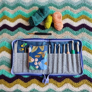 May include: A blue crochet hook case with a zipper closure. The case is open and contains a variety of crochet hooks, as well as a small pouch with a floral pattern. The case is sitting on a crocheted blanket with a wavy pattern in shades of blue, green, and brown.