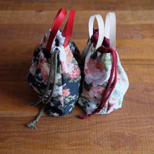 May include: Two fabric drawstring bags with floral patterns. One bag is black with white and pink flowers and has red handles. The other bag is white with pink and green flowers and has white handles.