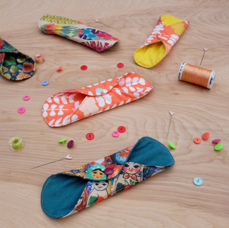May include: Four reusable cloth menstrual pads in various colours and patterns. The pads are folded and laid out on a wooden surface. The pads are made of fabric with different designs, including floral, geometric, and a Russian doll pattern. There are also sewing supplies scattered around the pads, including spools of thread, buttons, and pins.