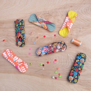 May include: A collection of colourful fabric menstrual pads with various patterns, including floral, geometric, and vintage illustrations. The pads are folded and arranged on a wooden surface.