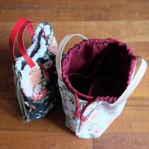 May include: Two floral fabric drawstring bags with different colored linings. One bag has a red drawstring and white lining, the other has a white drawstring and a burgundy lining.