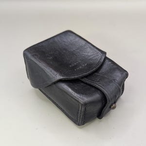 Könnte beinhalten: Ein kleiner, rechteckiger schwarzer Lederkoffer mit Klappenverschluss. Der Koffer hat eine strukturierte Oberfläche und ein Metallnietendetail an der Seite. Der Koffer scheint Vintage und abgenutzt zu sein.
