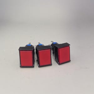 Op de afbeelding: Drie rode rechthoekige drukknopschakelaars met zwarte frames en zilveren connectoren. De schakelaars staan tegen een effen, lichtgrijze achtergrond. De knoppen zijn gelijkmatig verdeeld.
