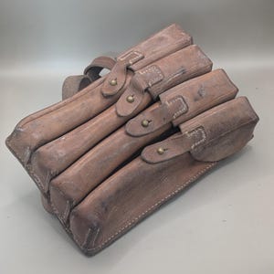 May include: Four brown leather pouches are stacked together. Each pouch has a flap secured by a strap and a brass button. The leather shows signs of wear and age, with visible stitching.