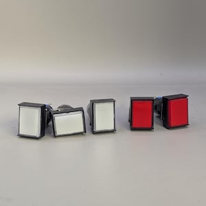 Puede incluir: Un surtido de pulsadores eléctricos rectangulares. Tres botones tienen caras blancas y dos tienen caras rojas. Cada botón está encerrado en una carcasa negra.