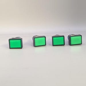 Könnte beinhalten: Vier grüne, rechteckige Drucktasten mit schwarzem Sockel sind horizontal vor einem weißen Hintergrund angeordnet. Die Tasten haben eine leuchtend grüne, beleuchtete Oberfläche. Dies sind wahrscheinlich elektrische Komponenten.