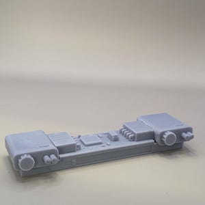 Op de afbeelding: Een lichtgrijs, rechthoekig elektronisch onderdeel met diverse verhoogde details, waaronder ronde knoppen en rechthoekige panelen. Het onderdeel heeft een complex, technisch ontwerp.