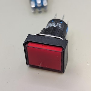 May include: A close-up of a red rectangular illuminated push button switch with a black frame. The switch has a black cylindrical body with silver pins and is set against a light background. Another switch is slightly out of focus in the background.
