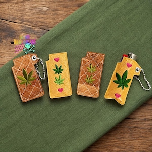 May include: Four rectangular lighter sleeves made of cork material with quilted stitching. Three sleeves feature embroidered green cannabis leaves and small pink hearts. One sleeve holds a silver lighter and includes a silver ball chain keychain.