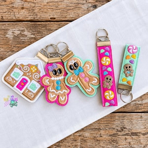 Può includere: Una collezione di portachiavi ricamati che include una casetta di pan di zenzero, un omino di pan di zenzero con fiocco viola, uno con fiocco blu e due portachiavi rettangolari da polso con motivi di caramelle in rosa e verde chiaro.