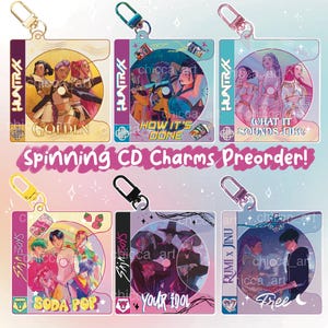 May include: A collection of spinning CD charms with colorful artwork and attached keychains. The charms feature various designs with text including "GOLDEN", "HOW IT'S DONE", "WHAT IT SOUNDS LIKE", "SODA POP", "YOUR IDOL", and "RUMI X JINU".
