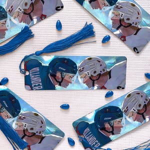 Könnte beinhalten: Lesezeichen mit Illustrationen von Eishockeyspielern, jeweils mit einer blauen Quaste und tropfenförmigen Perlen. Die Kunstwerke zeigen Spieler mit Helmen, wobei der Name "VOLLANDER" auf dem Trikot eines Spielers zu sehen ist. Die Farbpalette umfasst Blau-, Weiß- und Rottöne.