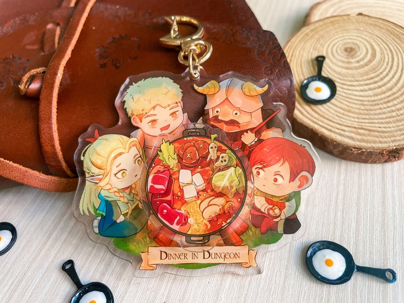 May include: A colorful acrylic charm featuring four cartoon characters enjoying a hot pot meal with the text "Dinner in Dungeon".
