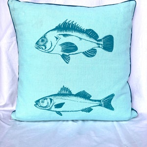 Könnte beinhalten: Ein hellblaues Kissen mit einem blaugrünen Rand zeigt zwei Fischillustrationen. Der obere Fisch ist größer und hat ein detaillierteres Design. Der untere Fisch ist kleiner und hat ein einfacheres Design.