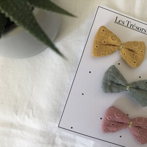 May include: Three fabric hair bows in different colors: mustard yellow, sage green, and dusty rose. Each bow has a gold accent in the center. The bows are displayed on a white card with the text "Les Tr&eacute;sors de Jaya".