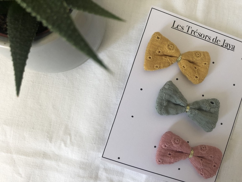 May include: Three fabric hair bows in different colors: mustard yellow, sage green, and dusty rose. Each bow has a gold accent in the center. The bows are displayed on a white card with the text "Les Tr&eacute;sors de Jaya".