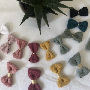 May include: A collection of handmade fabric bows in various colors, including white, pink, red, green, blue, and yellow. The bows are made with a delicate lace pattern and have a gold accent in the center.