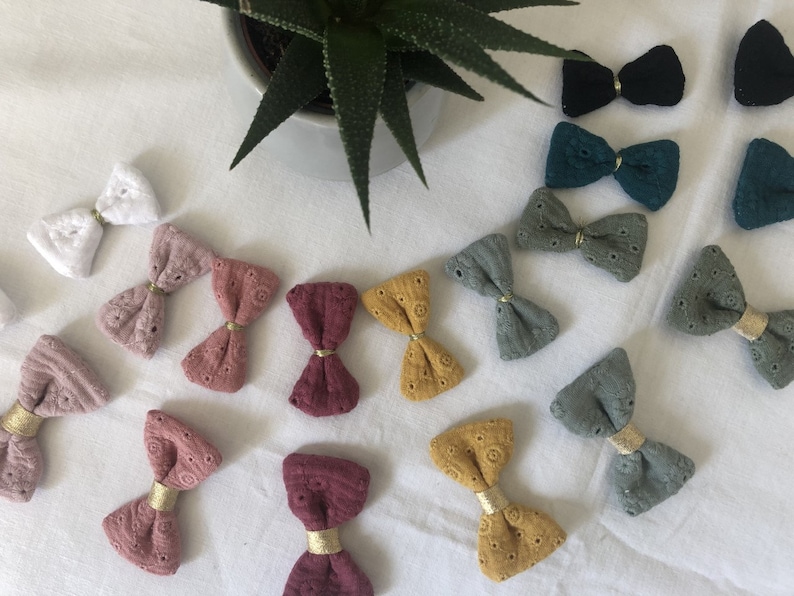 May include: A collection of handmade fabric bows in various colors, including white, pink, red, green, blue, and yellow. The bows are made with a delicate lace pattern and have a gold accent in the center.