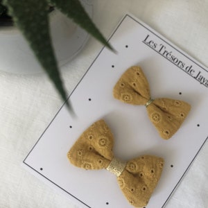 May include: Two mustard yellow fabric hair bows with a gold trim. The bows are on a white card with the text "Les Tr&eacute;sors de Jaya".
