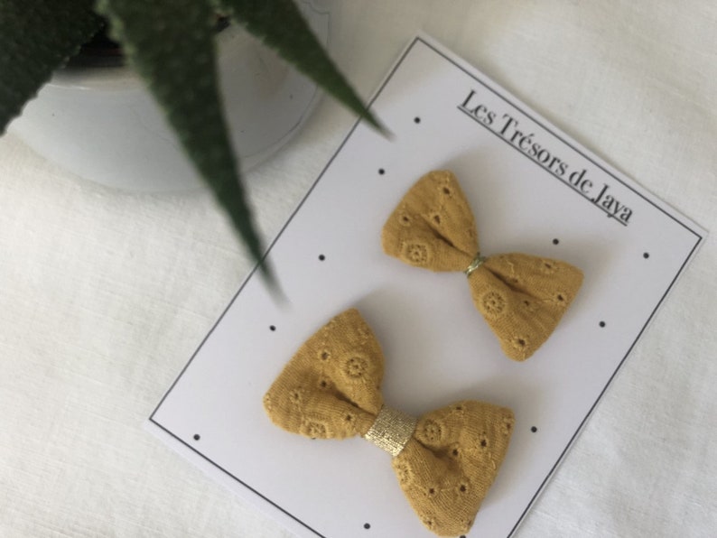 May include: Two mustard yellow fabric hair bows with a gold trim. The bows are on a white card with the text "Les Tr&eacute;sors de Jaya".