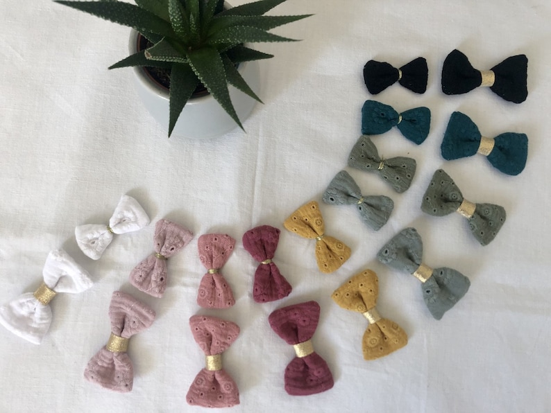 May include: Assortment of handmade fabric hair bows in various colors and patterns, including white, pink, green, and yellow. The bows are made with a delicate lace fabric and feature gold accents.