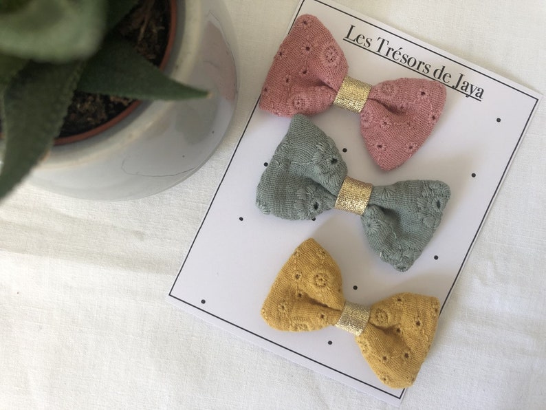 May include: Three fabric hair bows with gold trim. The bows are pink, green, and yellow. The bows are displayed on a white card with the text "Les Tr&eacute;sors de Java".