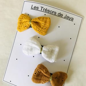 May include: Three fabric hair bows on a white card with black dots. The bows are in shades of yellow, white, and brown. The card has the text "Les Tr&eacute;sors de Jaya."