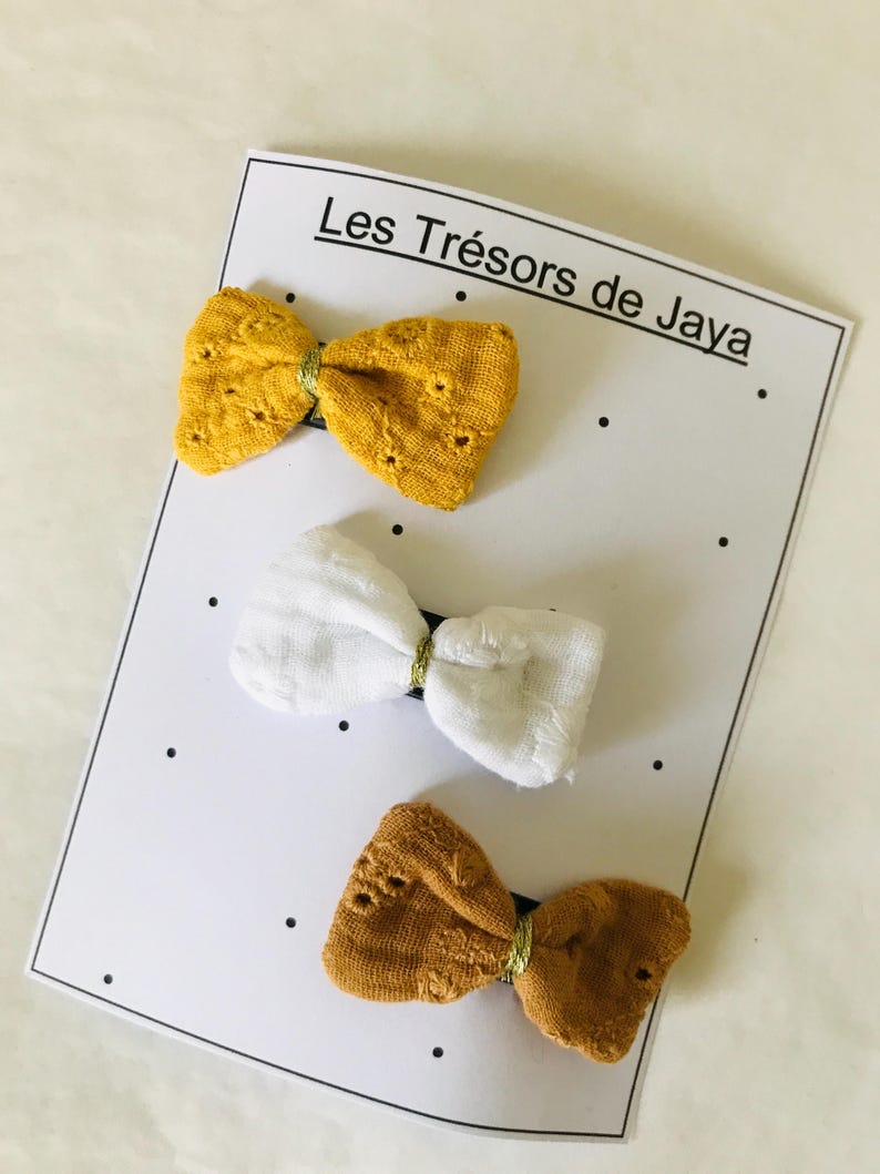 May include: Three fabric hair bows on a white card with black dots. The bows are in shades of yellow, white, and brown. The card has the text "Les Tr&eacute;sors de Jaya."