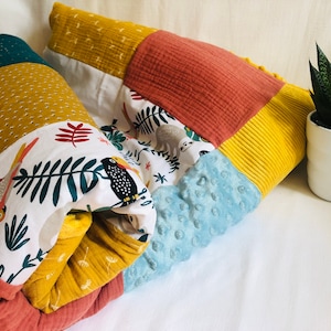 Peut inclure: Une couverture patchwork aux couleurs vives, avec des tissus jaunes, turquoise, corail et bleus. Elle présente un motif de toucans et de feuilles, un motif à pois et un tissu bleu texturé. Un petit pot de plante est à côté.
