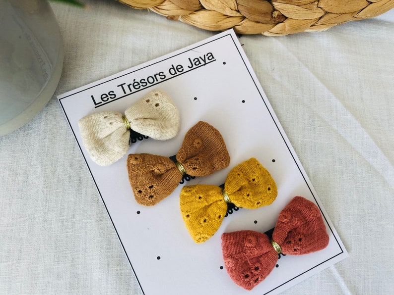 May include: Four fabric hair bows in different colors: cream, brown, yellow, and red. The bows are displayed on a white card with the text "Les Tr&eacute;sors de Jaya".