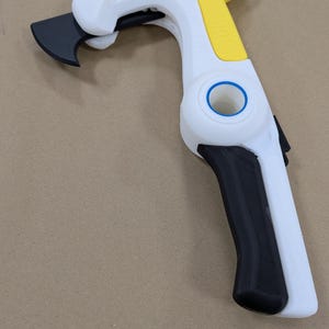 Puede incluir: Una herramienta manual con marco blanco, mango superior amarillo y empuñadura negra. La herramienta incluye una hoja negra curva en la parte frontal, una articulación circular central con un anillo azul y una sección amarilla texturizada.