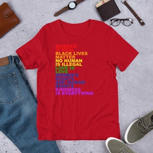 May include: Red t-shirt with a rainbow-colored message that reads "Science is real, Black Lives Matter, No Human is Illegal, Love is Love, Women's Rights are Human Rights, Kindness is Everything."
