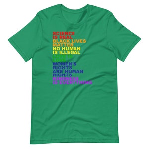 May include: A green t-shirt with a rainbow-colored message that reads "Science is real, Black Lives Matter, No Human is Illegal, Love is Love, Women's Rights are Human Rights, Kindness is Everything."