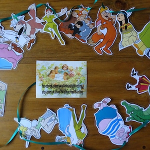 Puede incluir: Una cuerda verde con recortes de papel de personajes de Peter Pan, incluyendo Peter Pan, el Capitán Garfio, Campanita y Wendy. Los personajes están todos mirando en diferentes direcciones y están dispuestos en un círculo. La cuerda está sobre una superficie de madera.