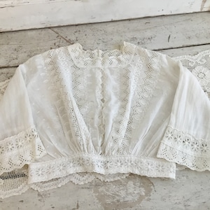 May include: A white, vintage-style blouse with intricate eyelet and lace detailing. The blouse features a square neckline, long sleeves with flared cuffs, and a gathered waist. The delicate fabric and floral patterns create a romantic, feminine aesthetic.