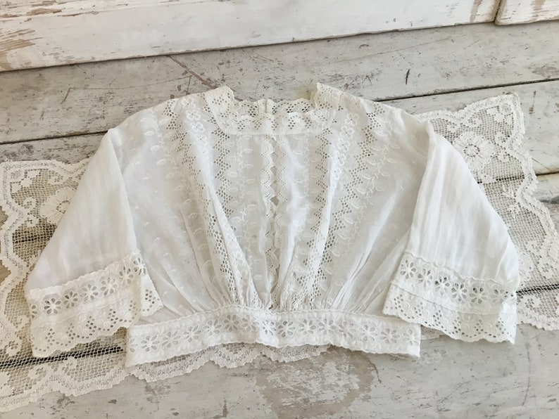 May include: A white, vintage-style blouse with intricate eyelet and lace detailing. The blouse features a square neckline, long sleeves with flared cuffs, and a gathered waist. The delicate fabric and floral patterns create a romantic, feminine aesthetic.