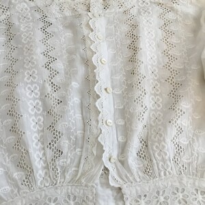 May include: White eyelet blouse with a high collar and button-down front. The blouse features intricate floral and leaf cutouts, scalloped edges, and button closures. The sleeves have a gathered cuff with floral embroidery.