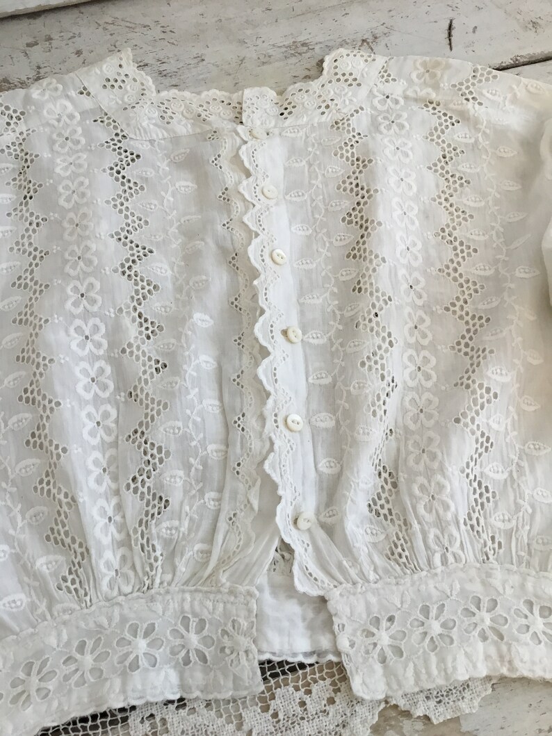 May include: White eyelet blouse with a high collar and button-down front. The blouse features intricate floral and leaf cutouts, scalloped edges, and button closures. The sleeves have a gathered cuff with floral embroidery.