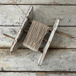 May include: A vintage wooden spool holder with a roll of natural twine. The holder has a ladder-like design with distressed white paint. The twine is beige and tightly wound. The background is a weathered, light-colored wooden surface.
