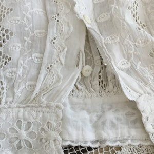 May include: Close-up of a white, vintage blouse with intricate floral embroidery and eyelet lace details. The blouse features button closures and a delicate, textured fabric, showcasing a timeless, romantic aesthetic.