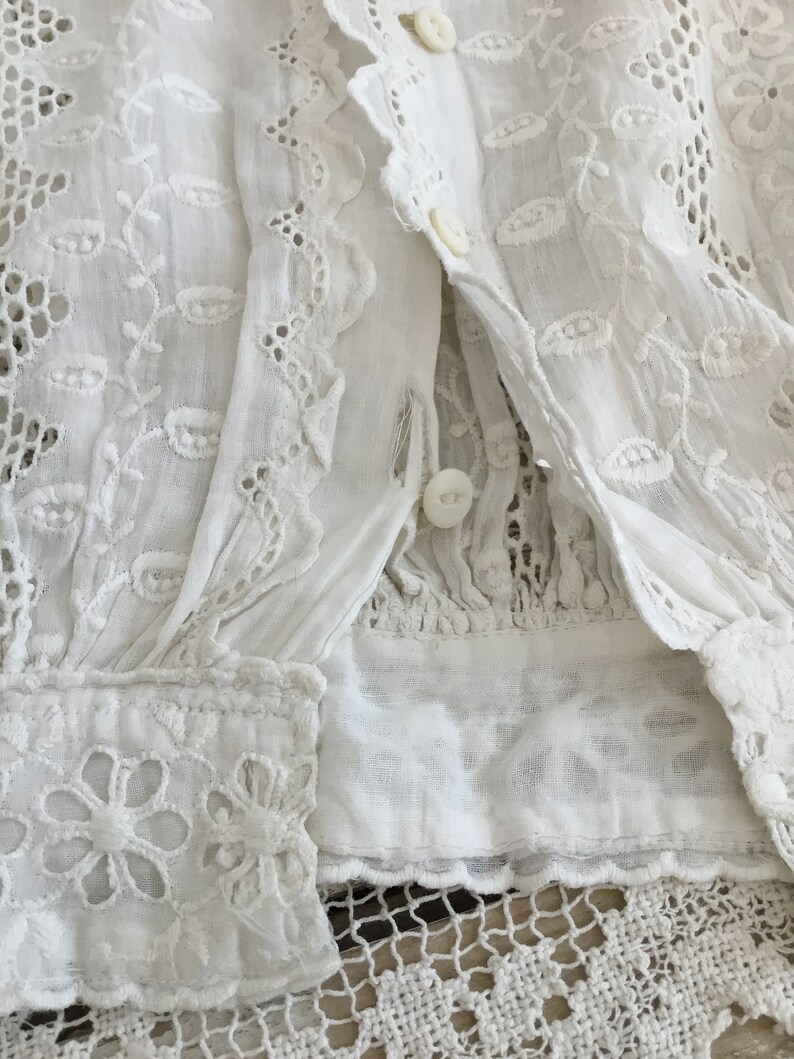 May include: Close-up of a white, vintage blouse with intricate floral embroidery and eyelet lace details. The blouse features button closures and a delicate, textured fabric, showcasing a timeless, romantic aesthetic.