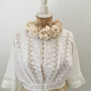 May include: A white, vintage-style blouse with intricate eyelet embroidery and short sleeves. The blouse features a fitted waist and a delicate floral trim. A cream-colored floral collar adorns the mannequin's neck, adding a touch of elegance to the garment.