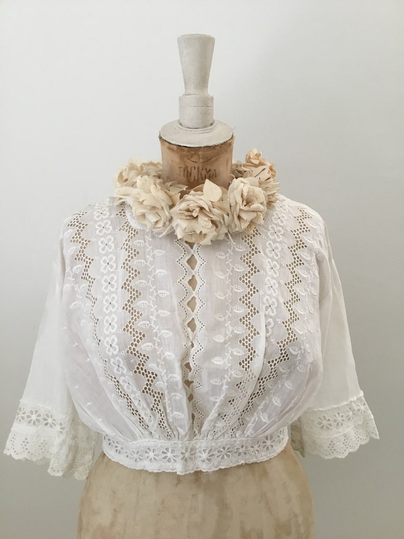 May include: A white, vintage-style blouse with intricate eyelet embroidery and short sleeves. The blouse features a fitted waist and a delicate floral trim. A cream-colored floral collar adorns the mannequin's neck, adding a touch of elegance to the garment.