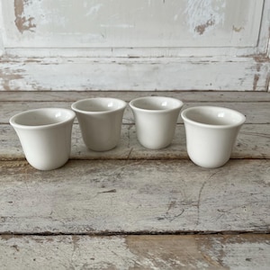 May include: Four small, white ceramic cups with a slightly flared rim. The cups are evenly spaced on a weathered, light-colored wooden surface. The background is a distressed white wooden panel.