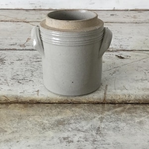May include: A light gray ceramic jar with a lid. The jar has two small handles and horizontal ridges around the body. The lid is a lighter tan color. The jar is sitting on a weathered, light-colored wooden surface.