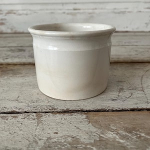 May include: A small, off-white ceramic jar with a slightly flared rim. The cylindrical body has a subtle horizontal band. The jar sits on a weathered, light-colored wooden surface, suggesting a vintage or rustic aesthetic. The jar is likely for kitchen or home decor.
