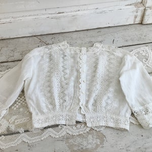 May include: A white, vintage-style blouse with long sleeves and intricate lace detailing. The blouse features a button-up back and delicate floral and geometric patterns. The sleeves and hem are trimmed with scalloped lace.